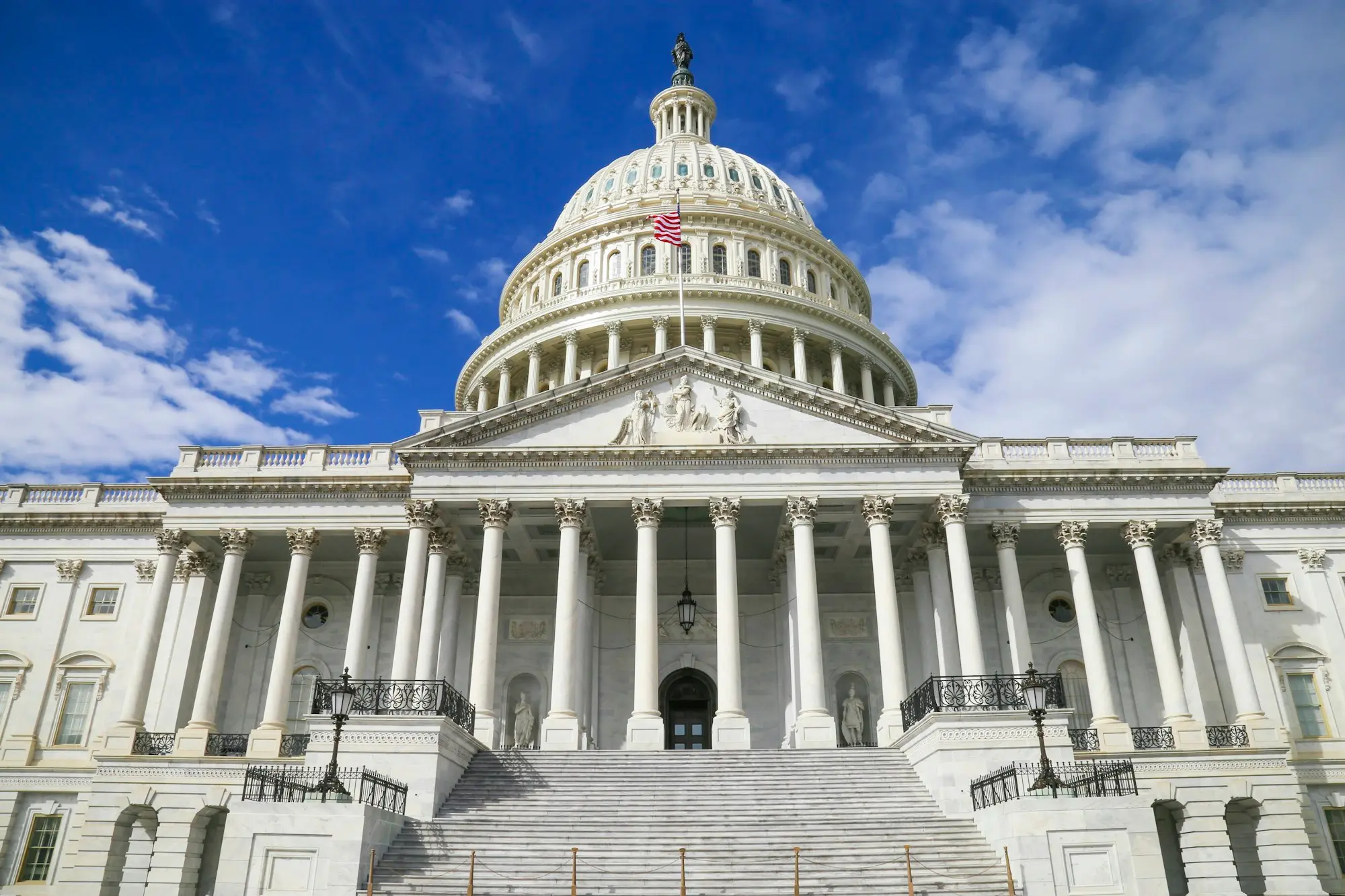 U.S. Capitol east front with its white dome and American flag under a bright blue sky with clouds