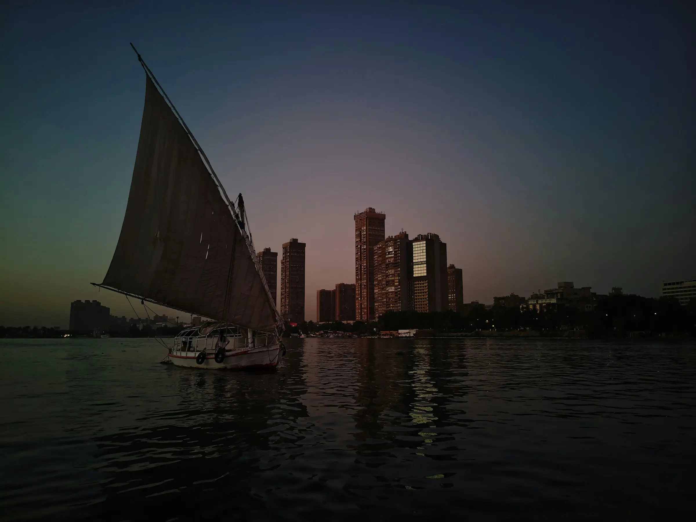 Felucca sailing on the Nile at dusk beside Cairo's riverside high-rises under a deep blue and violet sky