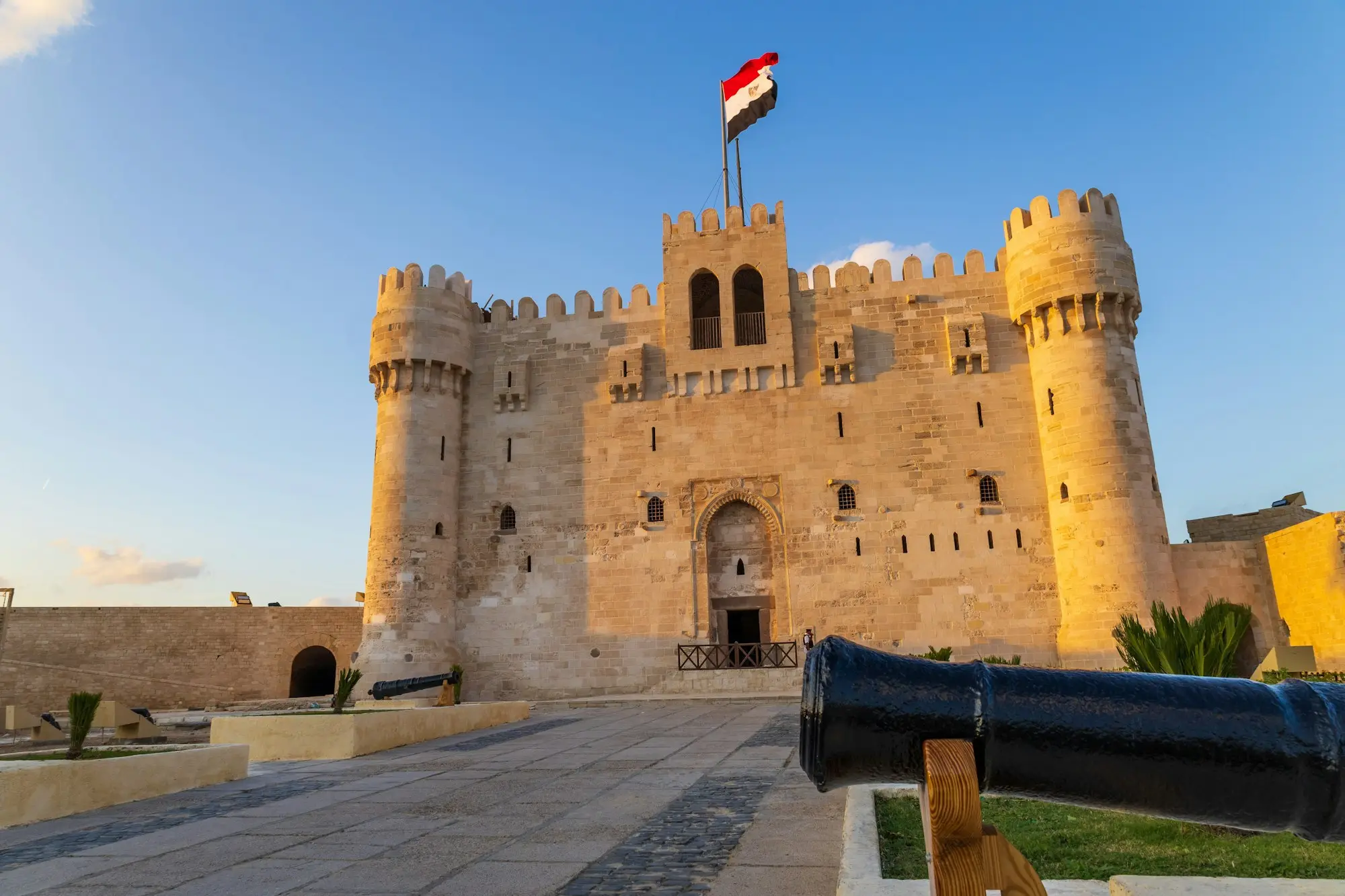 Citadel of Qaitbay in Alexandria glowing in golden light under a blue sky, Egyptian flag flying, cannon in foreground