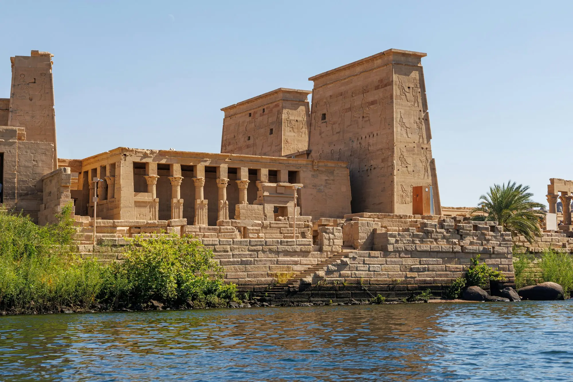 Philae Temple's carved pylons and colonnade rising above the Nile shoreline under a clear blue sky