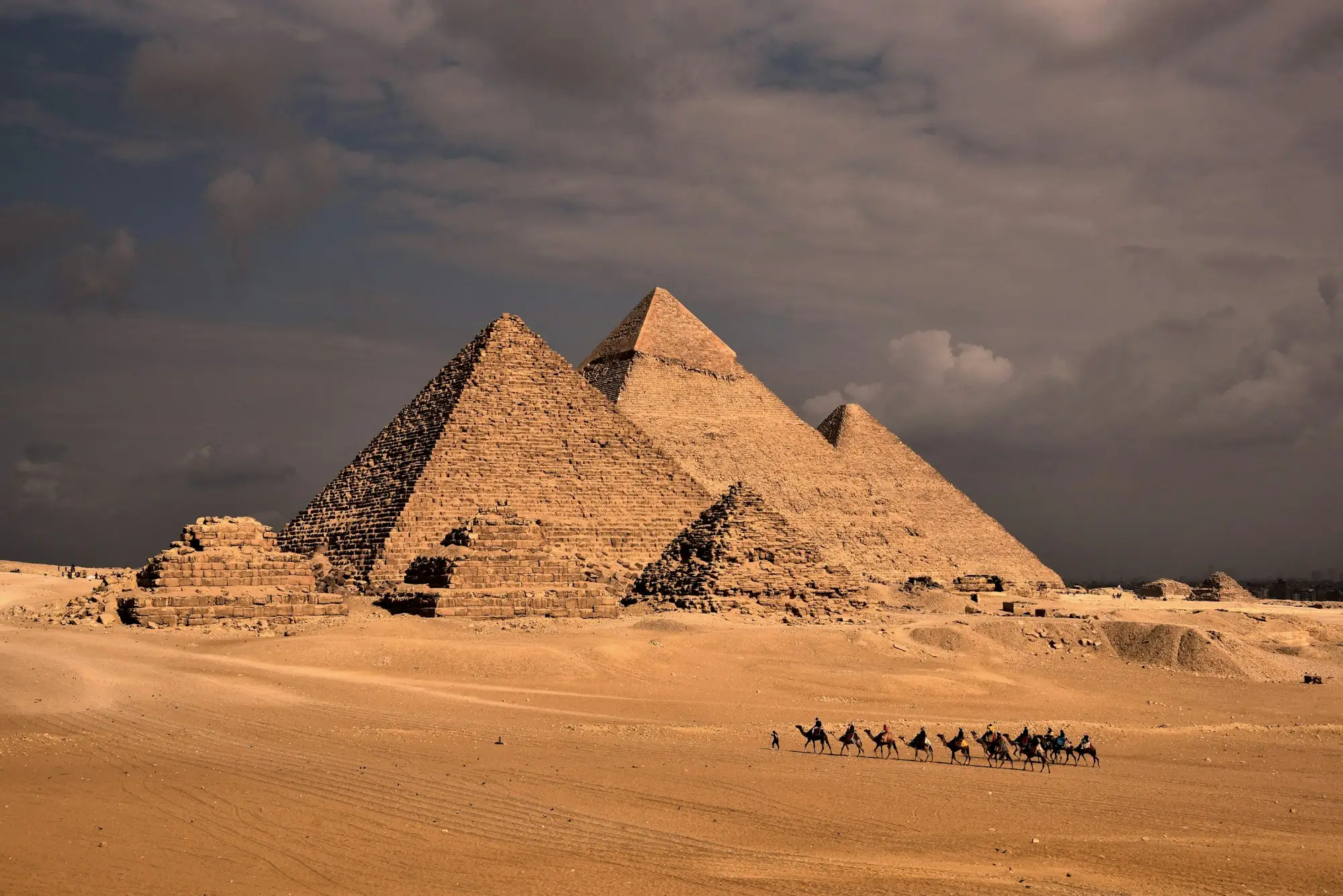 The Giza pyramids lined up in sunlight under a stormy sky, with a camel caravan crossing the sand in front