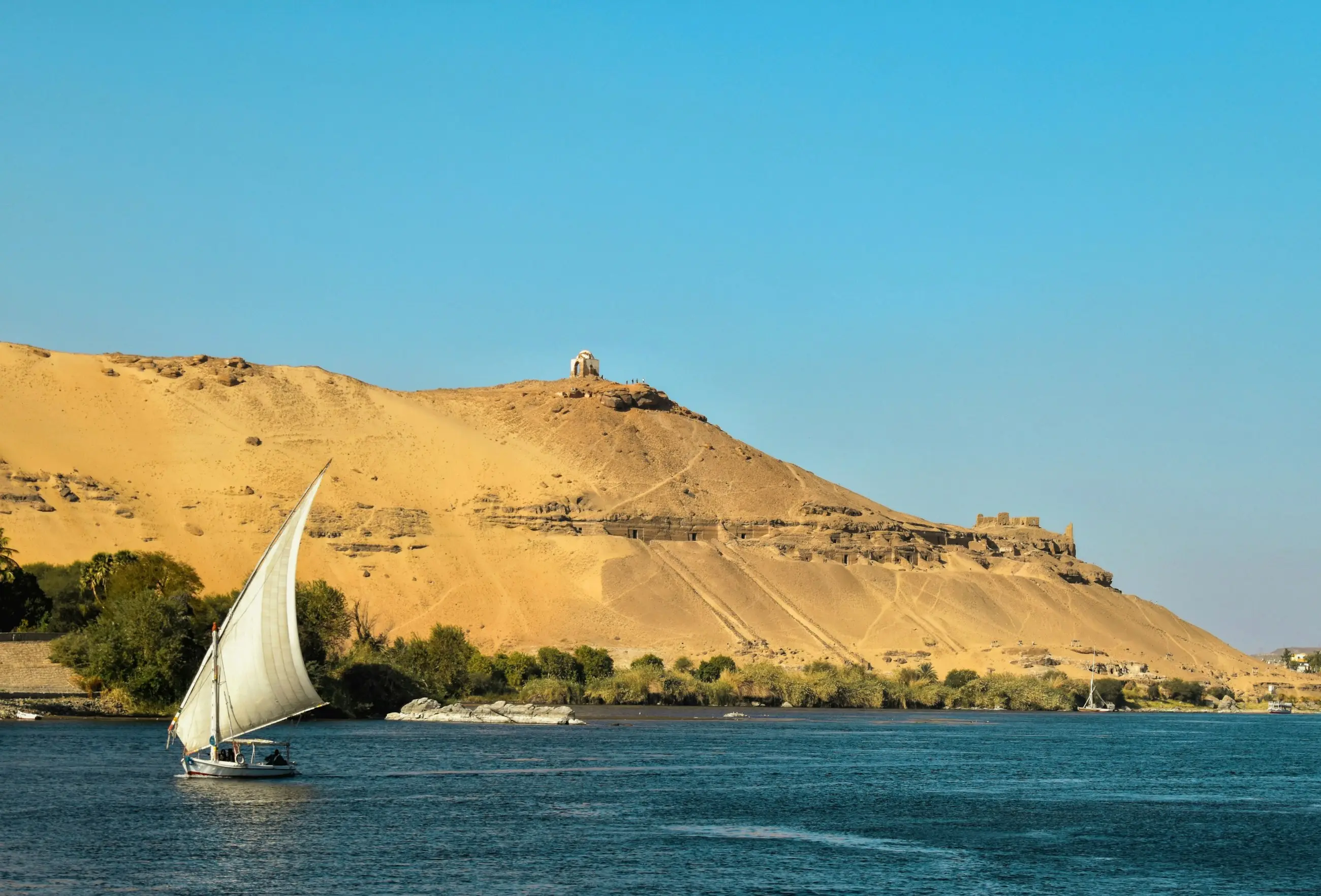 Felucca sailing on the Nile at Aswan below a golden desert hill with a domed shrine and rock-cut tombs