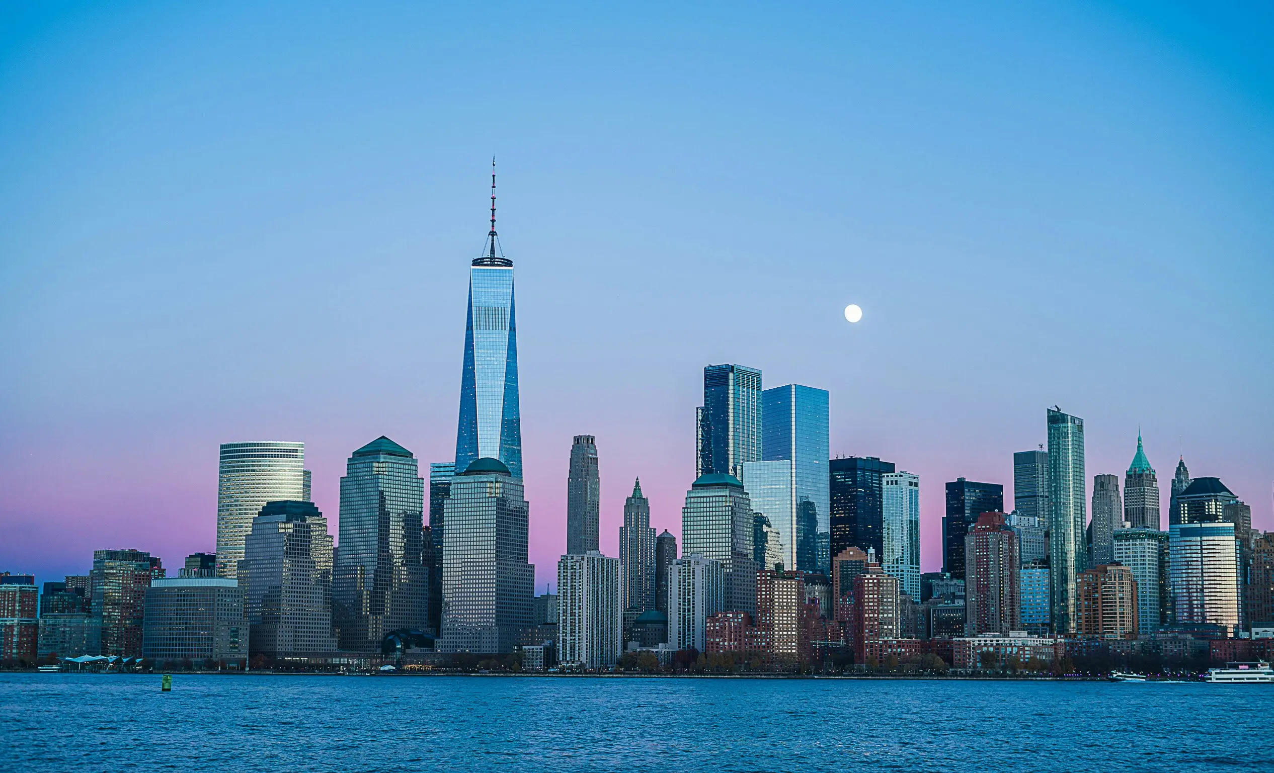Lower Manhattan skyline and One World Trade Center at blue hour across the Hudson River, with a full moon above