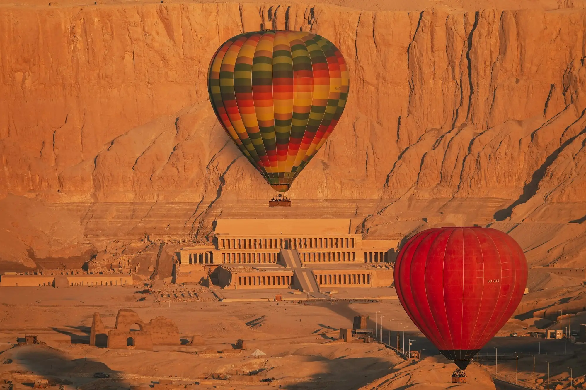 Two hot-air balloons drifting past the Temple of Hatshepsut and sunlit cliffs on Luxor's West Bank at sunrise