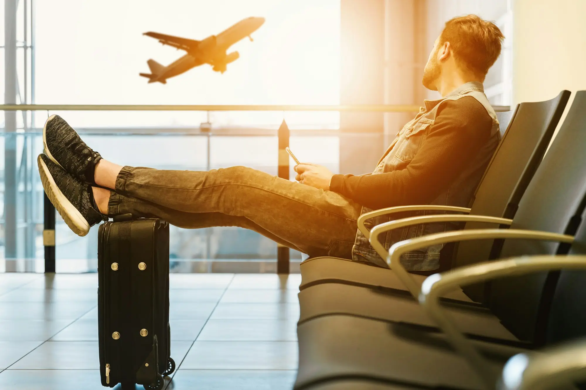Traveler with feet up on a black suitcase in an airport lounge, watching a plane take off through the window