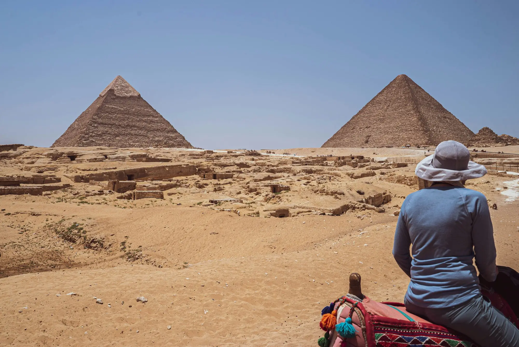 Traveler in a sun hat seen from behind on camelback, looking across the Giza plateau at two pyramids